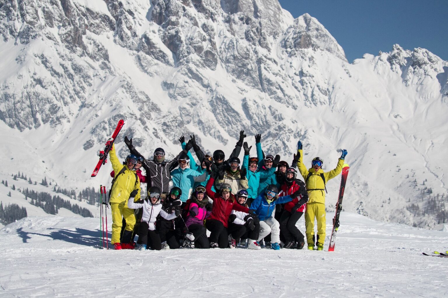 Eine  jubelnde  Gruppe aus 12 Skifahrerinnen und Skifahrern vor einen Bergkulisse in Skiausrüstung am Ende der Ausbildung.